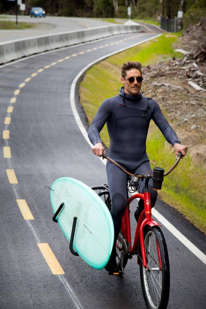Surfer in wetsuit pedals bike with surfboard on paved path